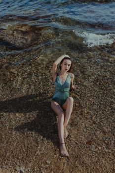 A woman in a swimsuit poses on a pebbly beach with waves gently crashing around her.