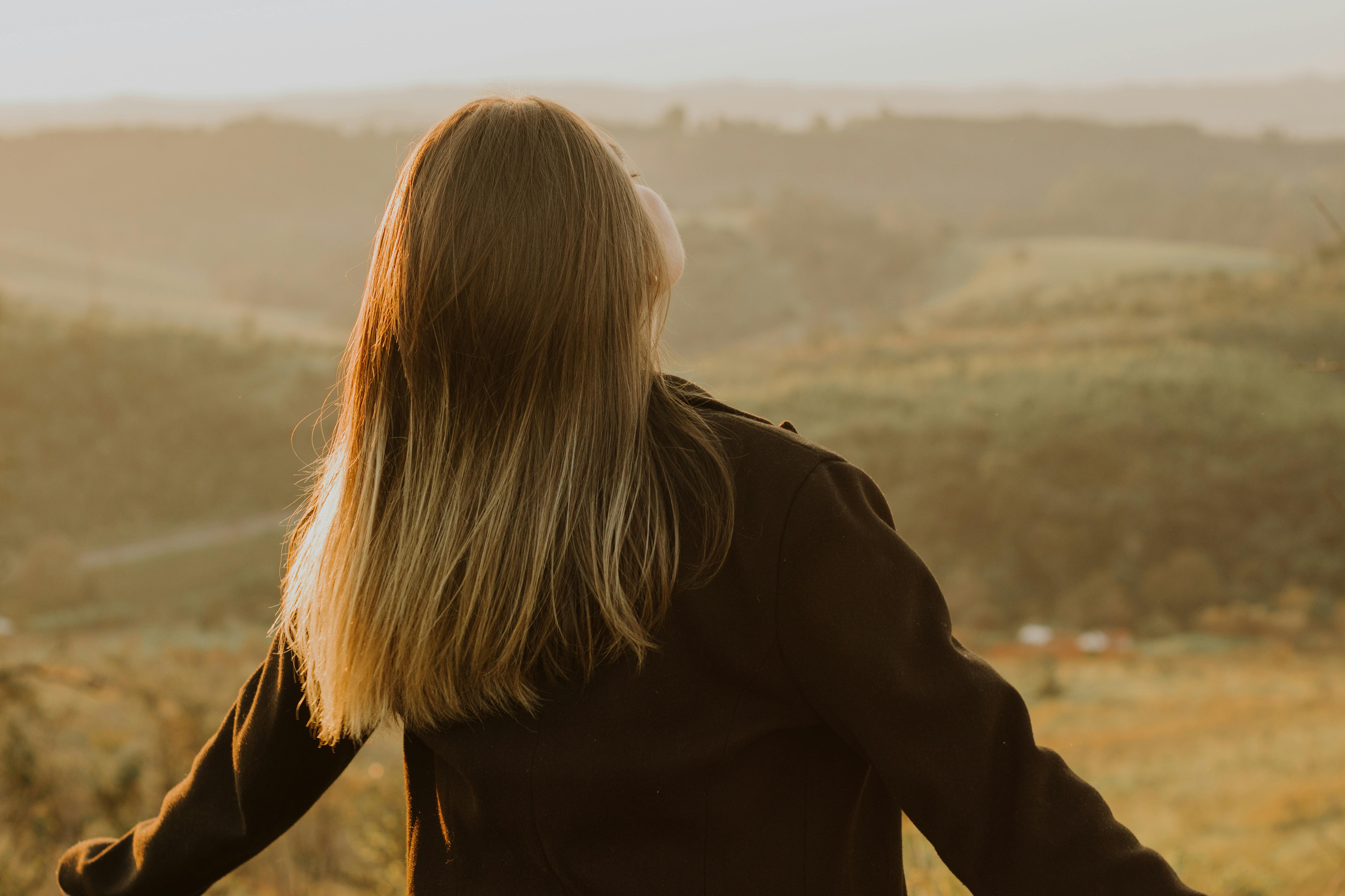 Back view of a woman with long hair standing in a scenic hillside at sunset, embracing nature.