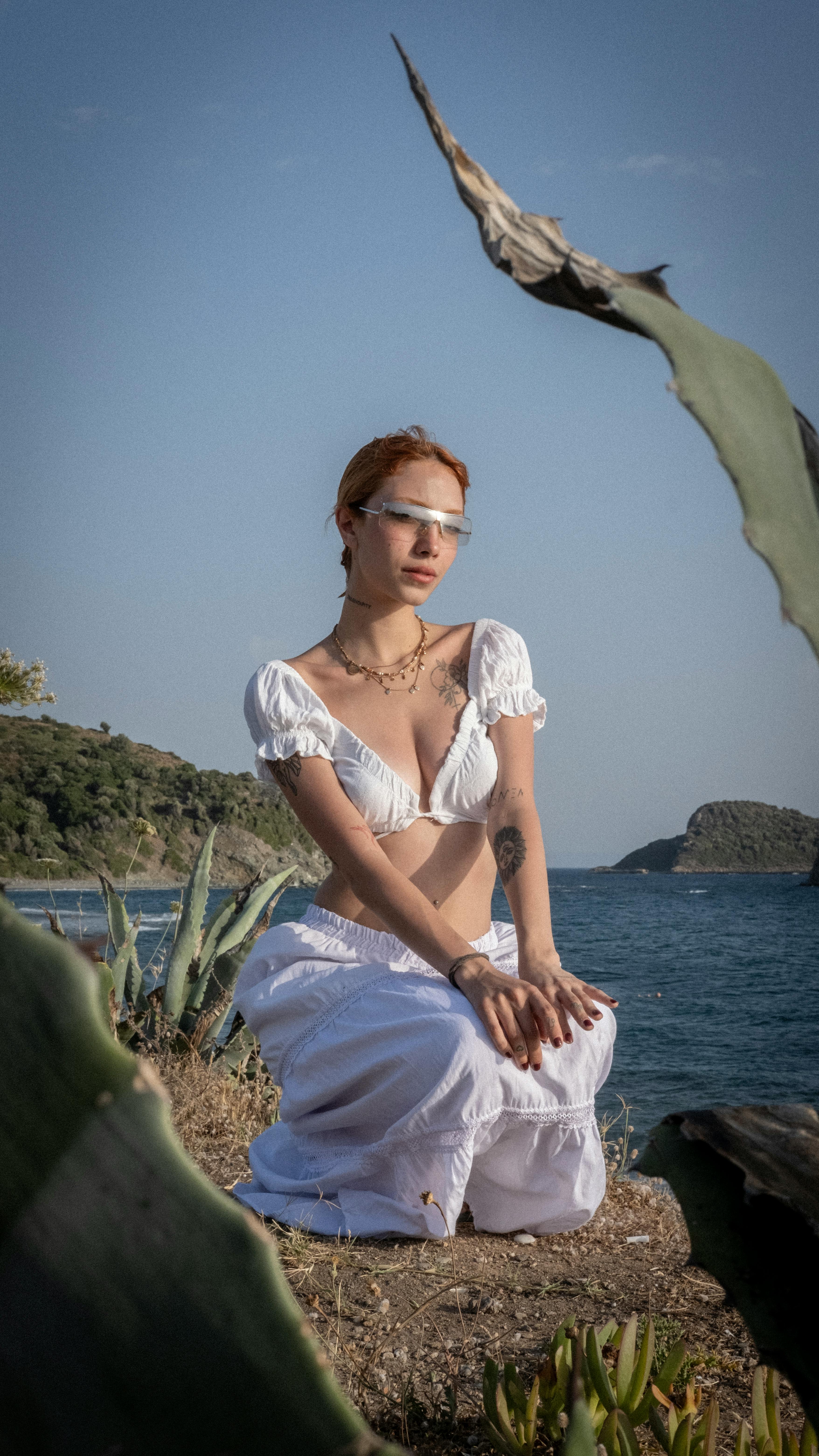 A stylish woman kneeling on the shore, wearing a white dress and sunglasses, with the ocean as a backdrop.