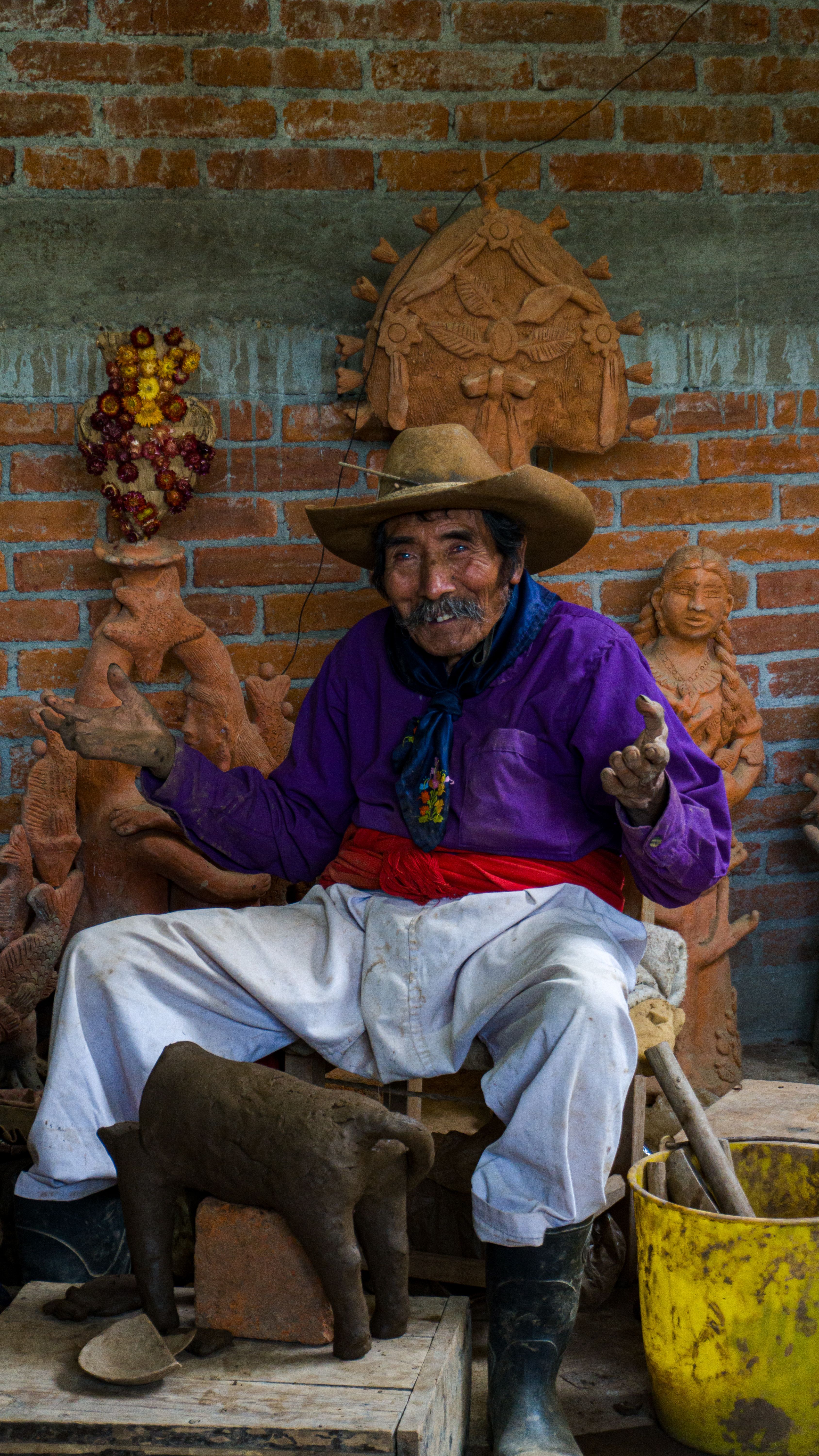 Fotomural Un artesano anciano con camisa púrpura esculpiendo figuras de arcilla, sentado contra una pared de ladrillo rústica en México.