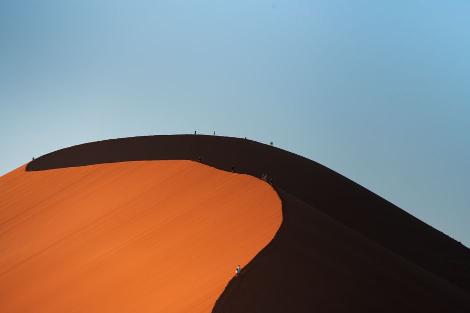 Silhouetted climbers ascend a massive orange desert dune beneath a clear blue sky.