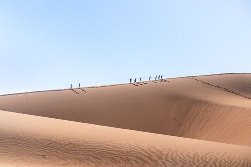 Free Group of people hiking across vast desert dunes with clear blue sky above, showcasing a serene and adventurous landscape. Stock Photo