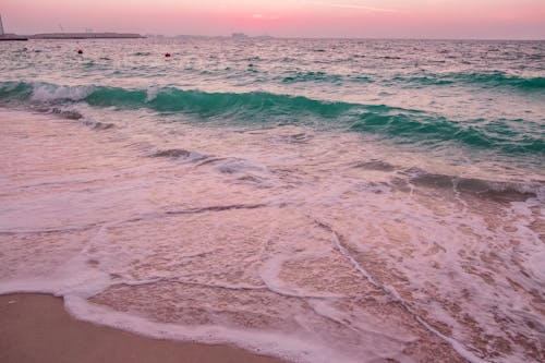 From above of foamy waves of turquoise ocean washing sandy beach against amazing pink sunset sky