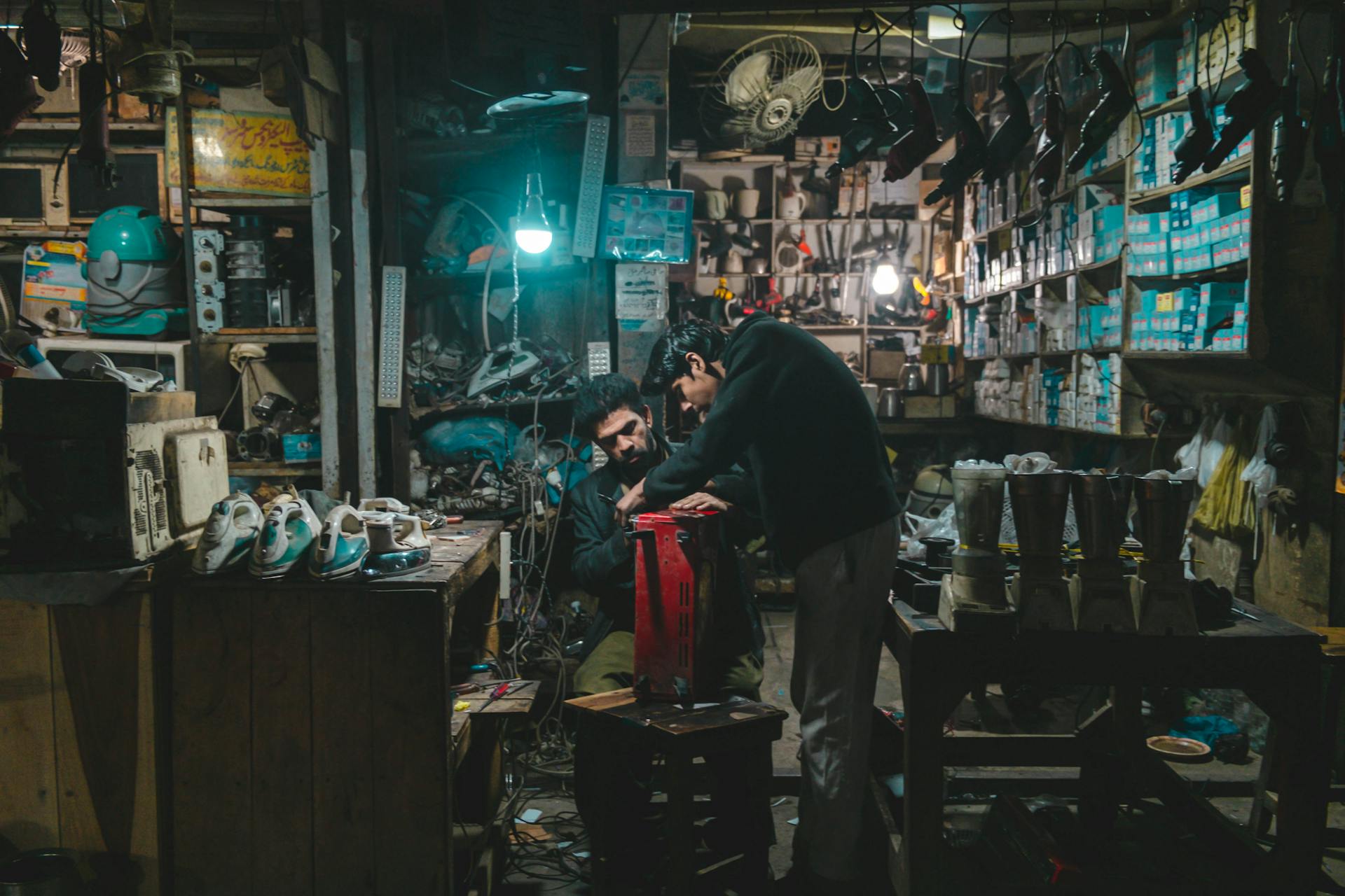 Two men working in a cluttered electronics repair shop in Faisalabad, Pakistan.