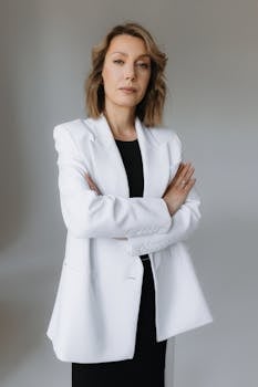 Confident businesswoman standing with arms crossed in a white suit, studio background.