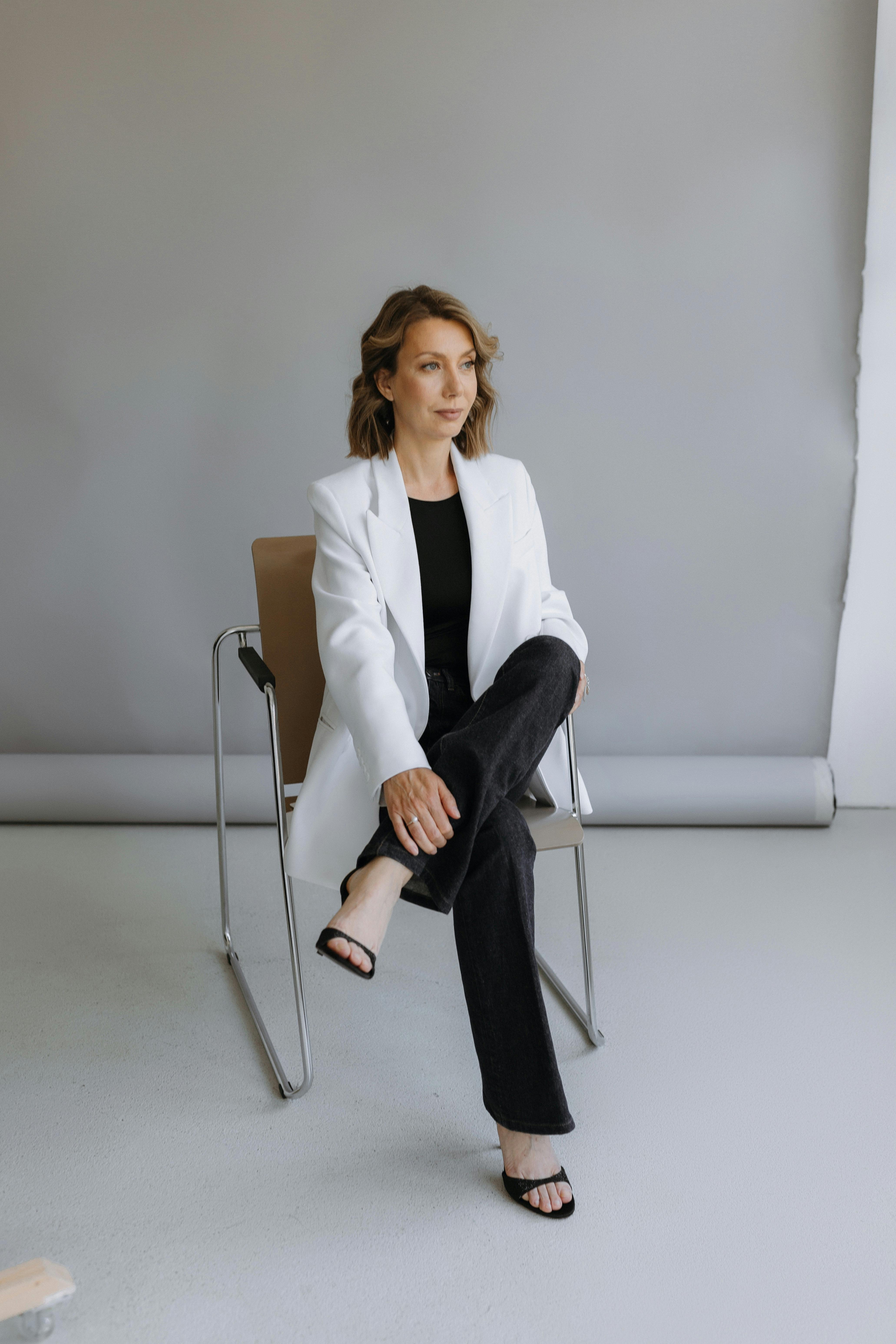Elegant woman with brown hair wearing a white blazer poses sitting on a chair in a studio setting.