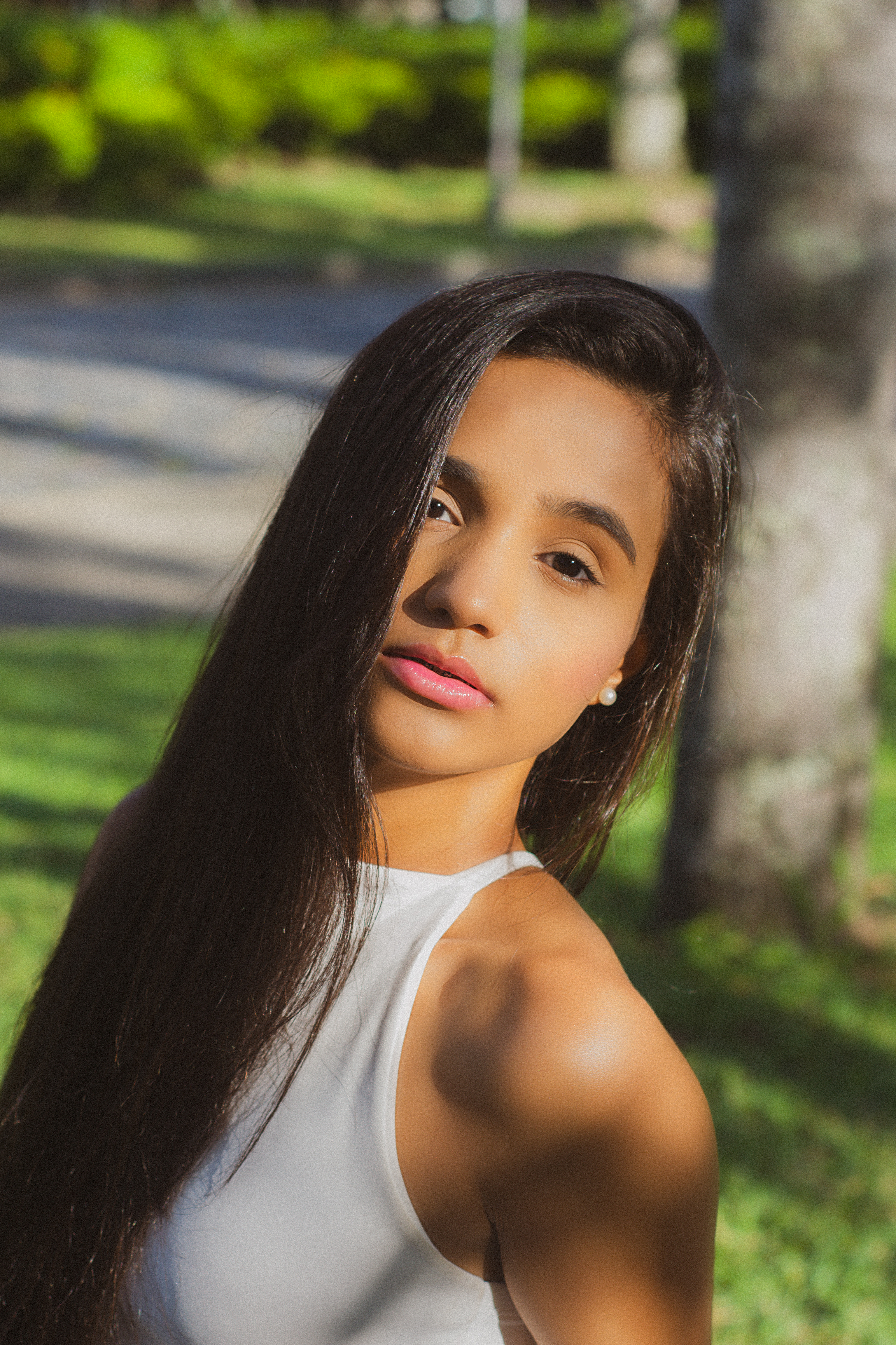 A young woman with long hair stands in a sunlit park, casting soft shadows.