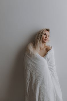 Elegant portrait of a blonde woman wrapped in a cozy white duvet, exuding calm and warmth indoors.