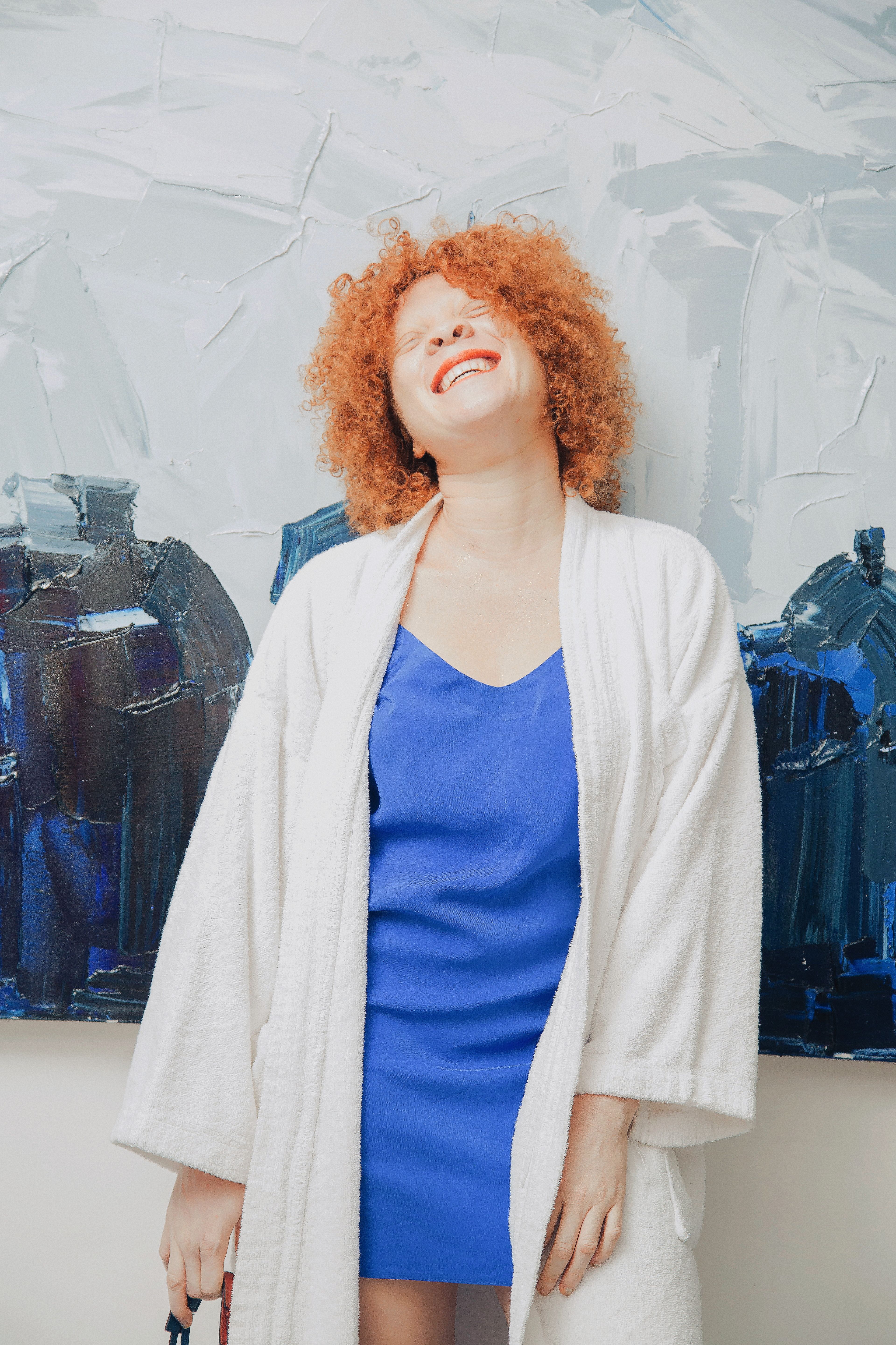 A joyful woman with curly orange hair smiling in a bathrobe against a modern background.