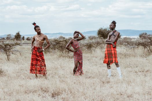 Three people in traditional tribal attire posing in a vast African grassland.