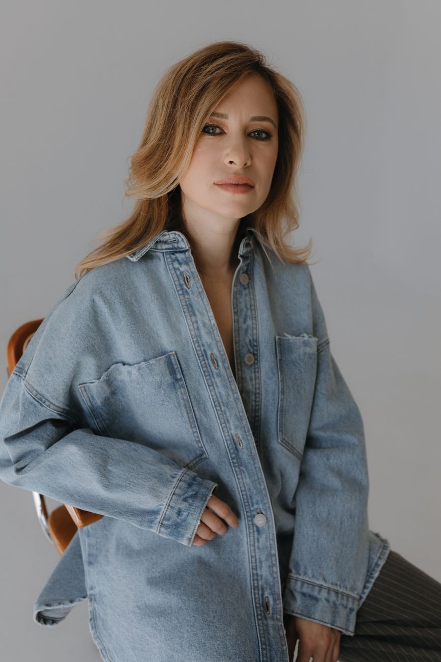 Elegant woman in a denim jacket sitting indoors with a minimalist background.