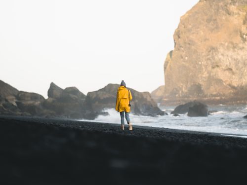 Free A woman in a yellow jacket walks along a tranquil, rocky beach, capturing a moment of peaceful solitude. Stock Photo