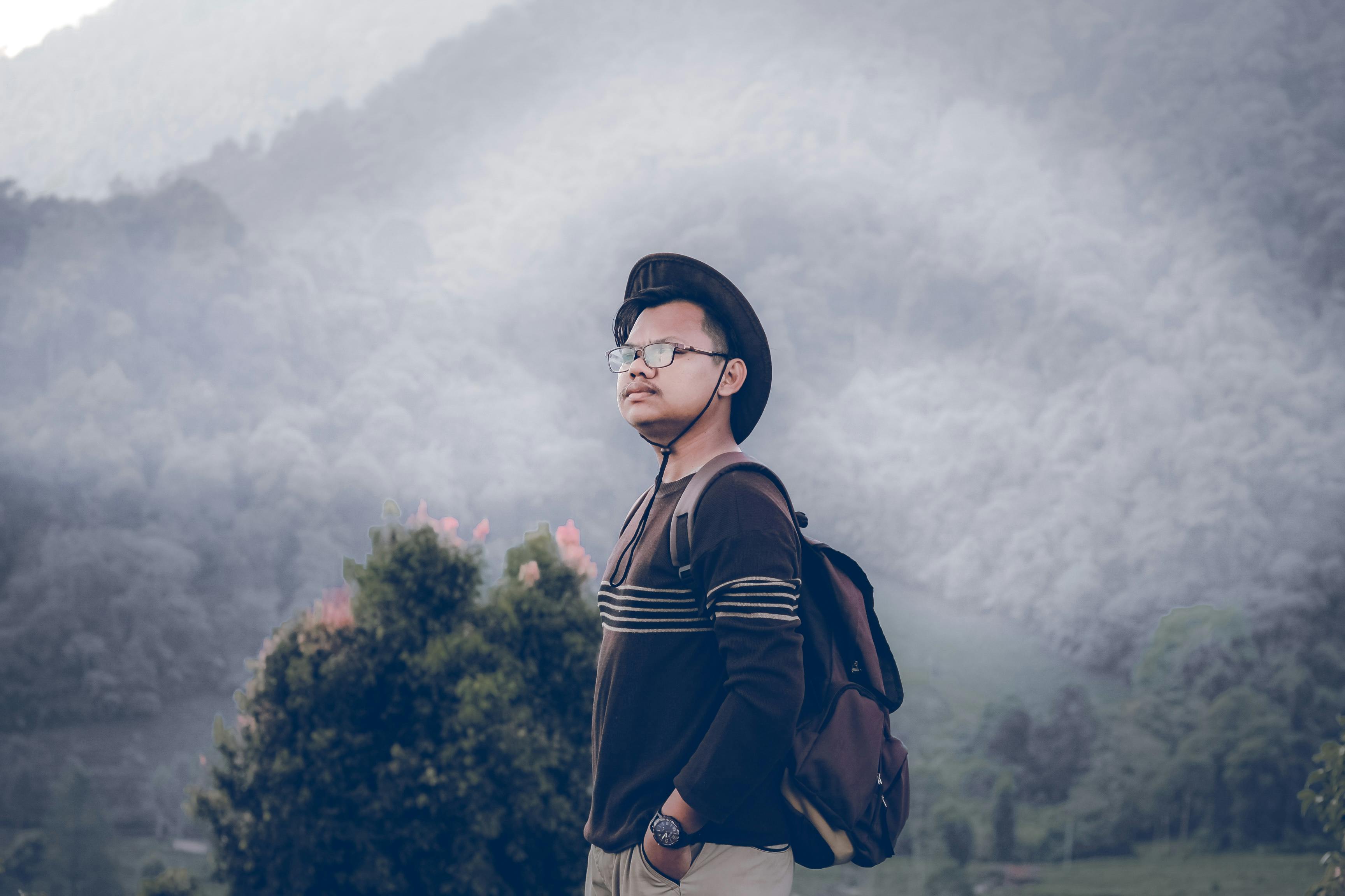 一名男子头戴帽子、身背背包，站在云雾缭绕的山间，尽显户外冒险精神