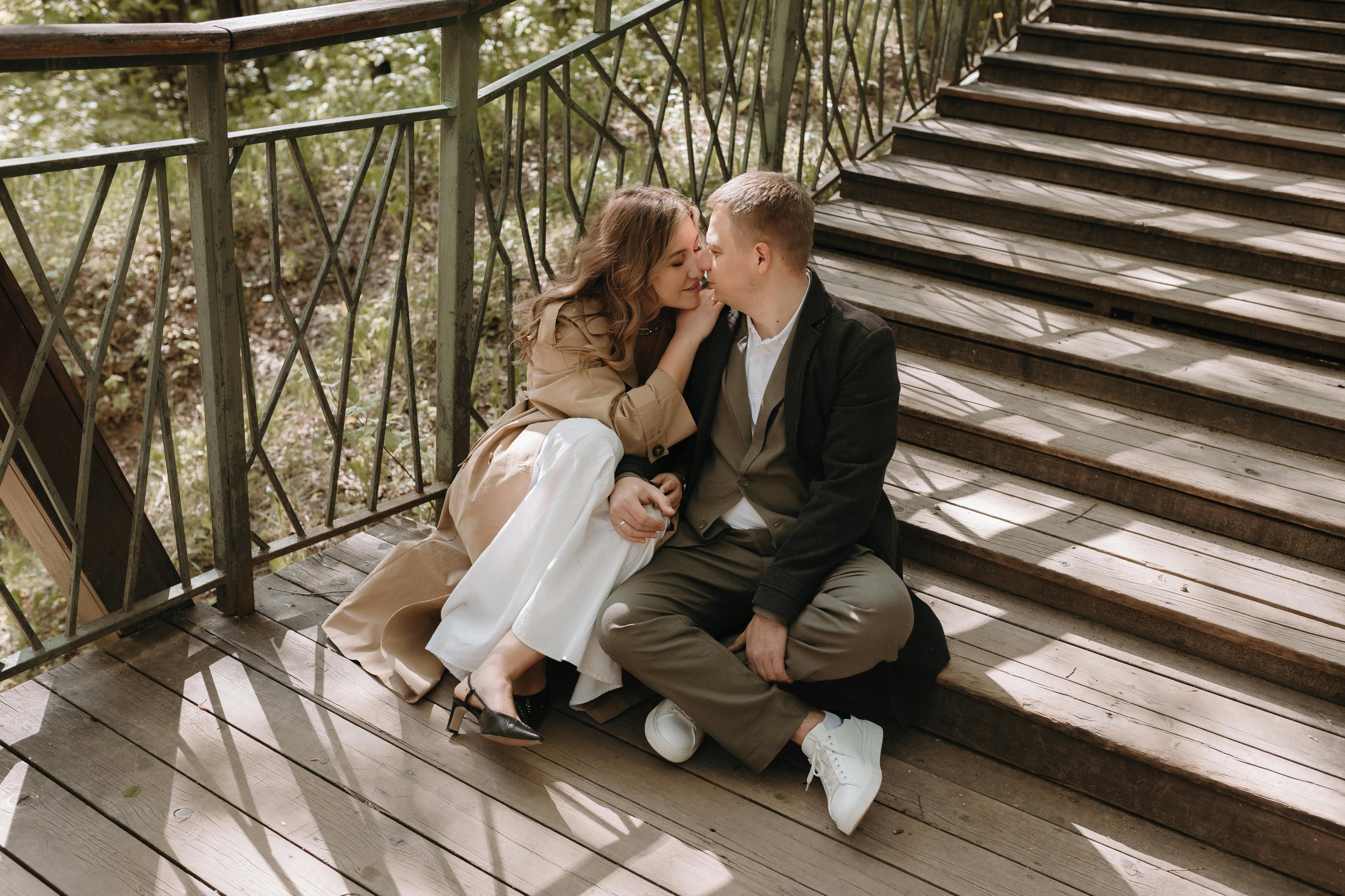 A couple sits affectionately on wooden stairs outdoors, enjoying a sunny day.