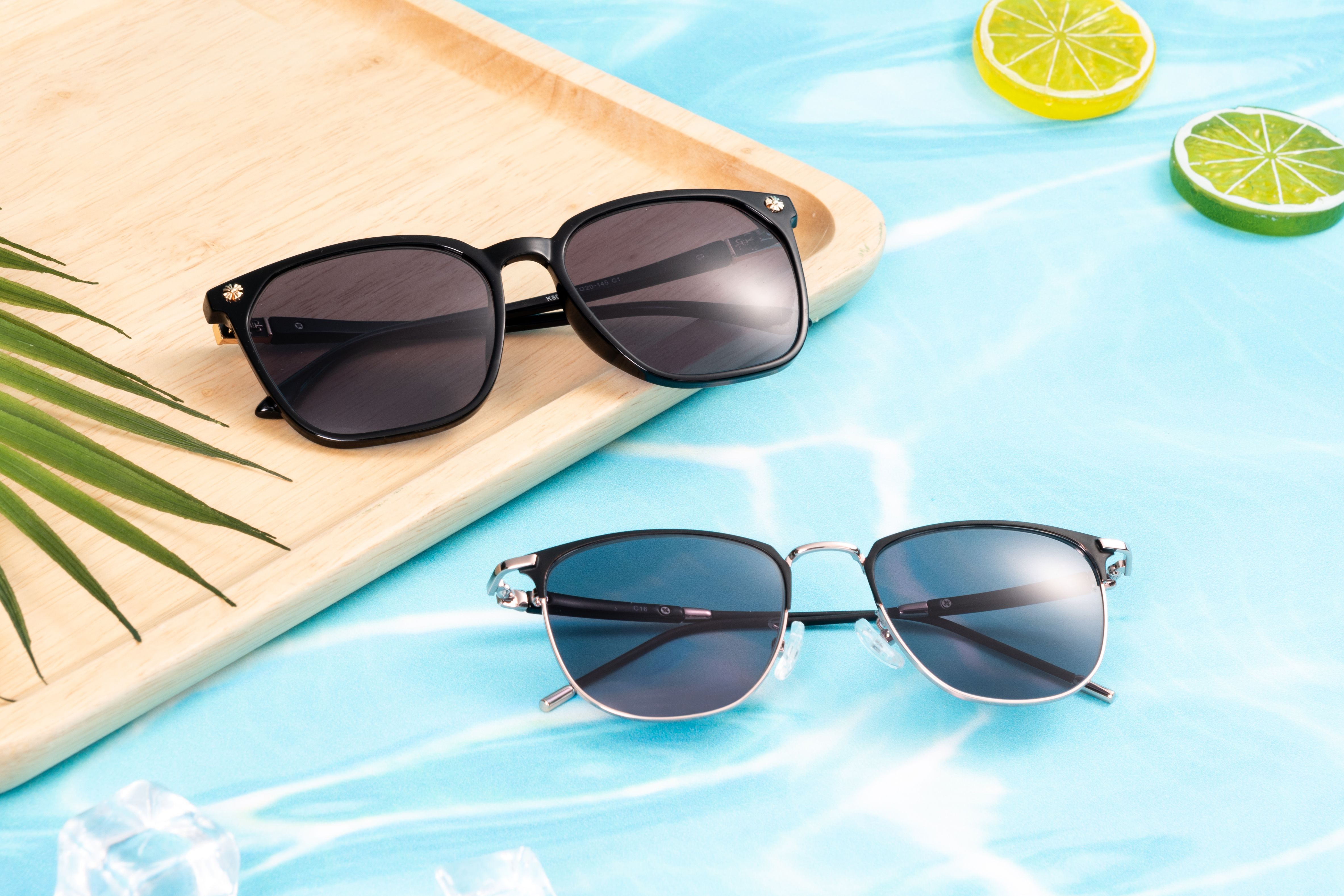 Elegant sunglasses on a wooden tray by a pool, evoking summer vibes with lime slices and palm leaf.