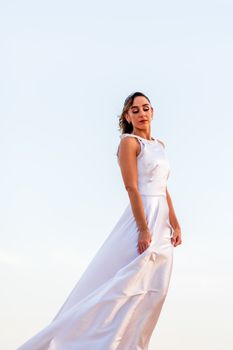 A graceful woman in a white dress poses outdoors against a blue sky, exuding elegance.