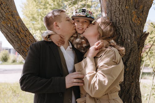 A man and woman kiss their child in front of a tree