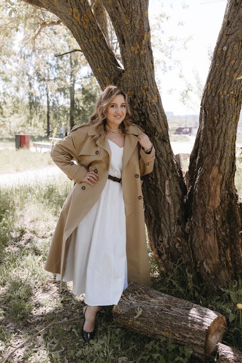 A woman in a white dress and trench coat posing near a tree