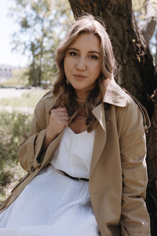 A woman in a white dress and trench coat sitting under a tree