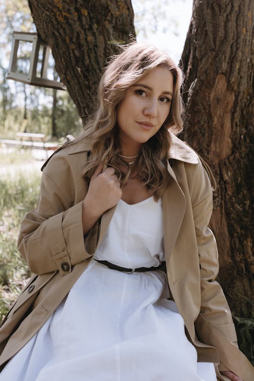 A woman in a white dress and trench coat sitting under a tree