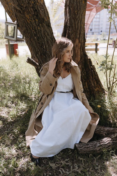 A woman in a white dress and trench coat sitting on a tree