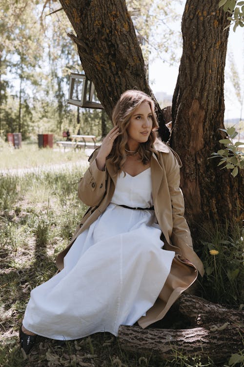 A woman in a white dress and trench coat sitting in a tree