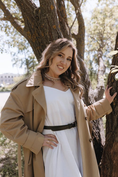 A woman in a white dress and trench coat posing near a tree