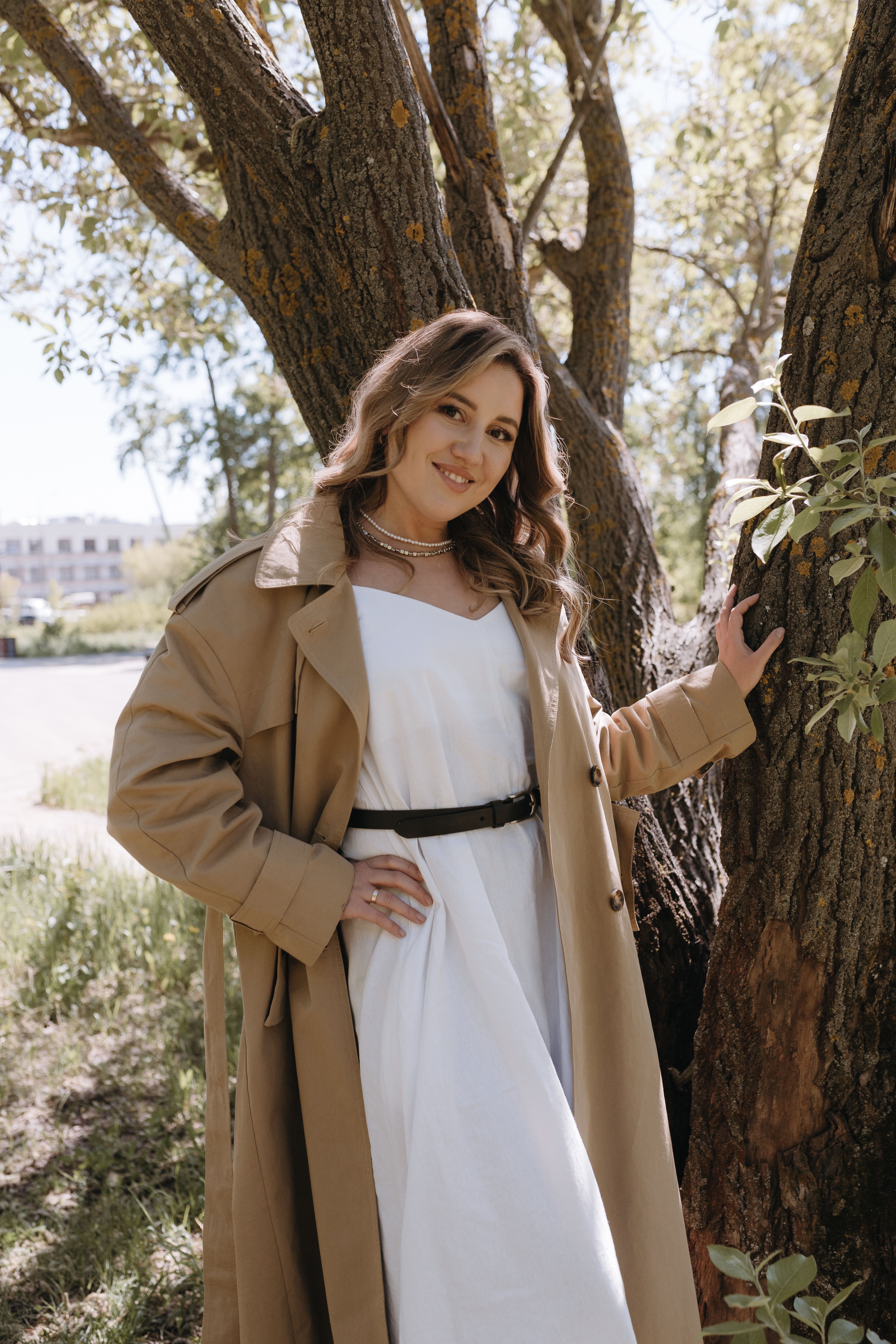 Woman in a white dress and trench coat posing near a tree