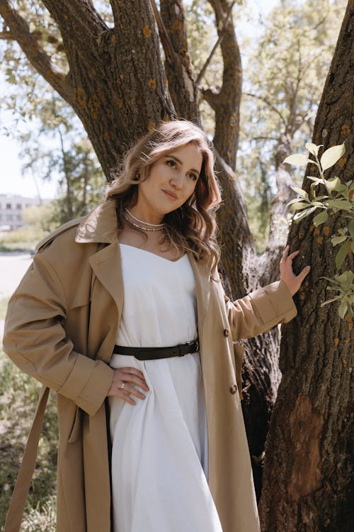 A woman in a white dress and trench coat posing near a tree