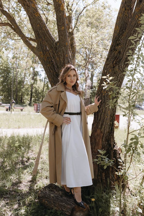 A woman in a white dress and trench coat standing next to a tree