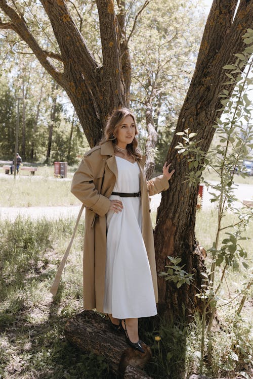 A woman in a white dress and a trench coat standing next to a tree