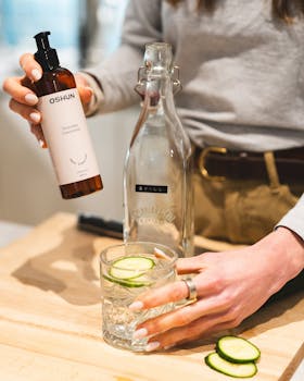 Woman preparing a cucumber-infused drink with skincare product in a cozy setting.