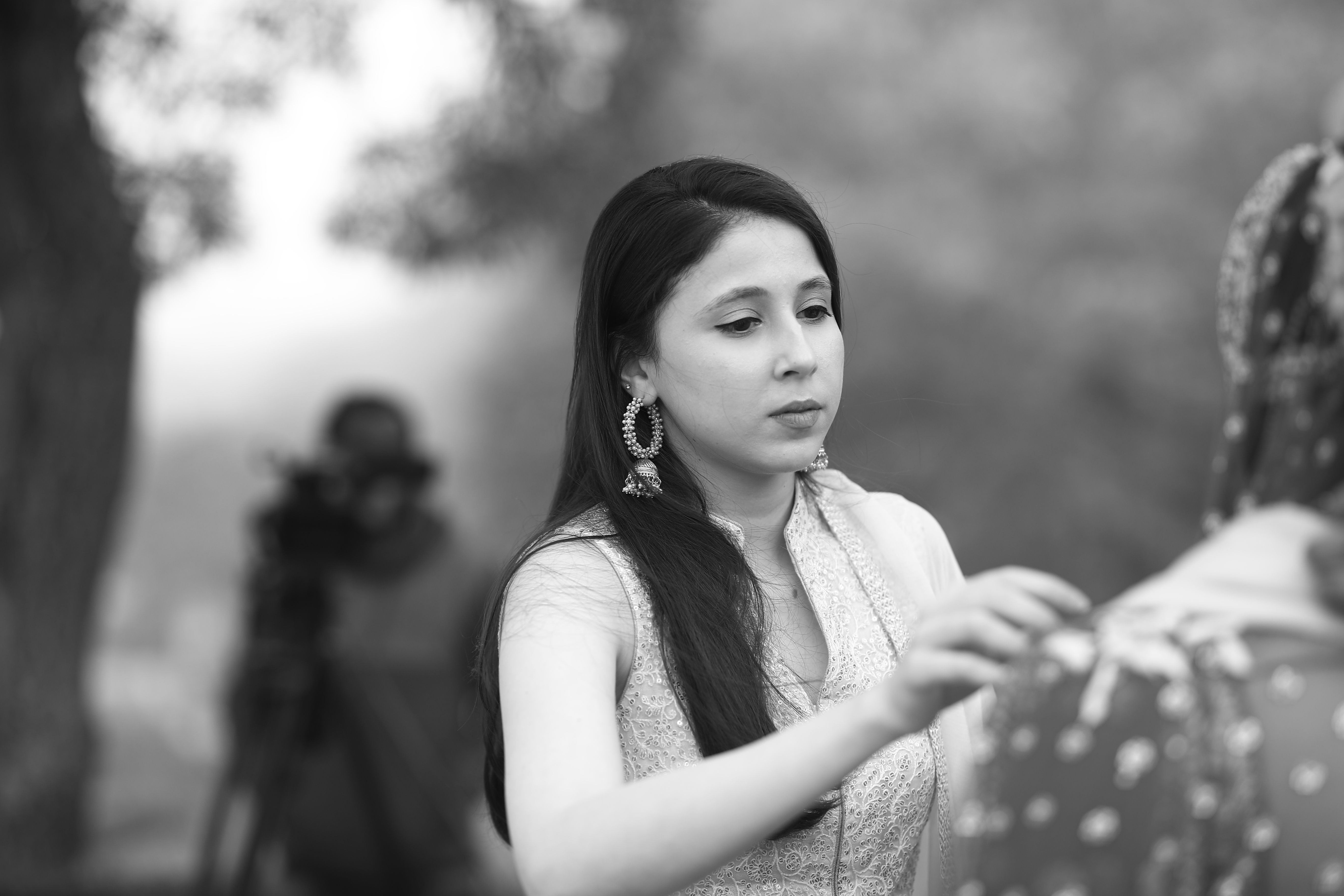An elegant woman adjusts a dress outdoors while a cameraman films in grayscale.