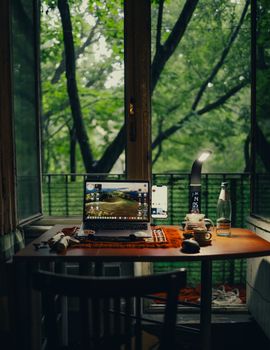 A peaceful home office setup on a balcony table, featuring a laptop and lamp, framed by a lush green outdoor view.
