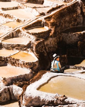 A woman in a hat sitting by the salt ponds of Maras, Peru, under the sun.