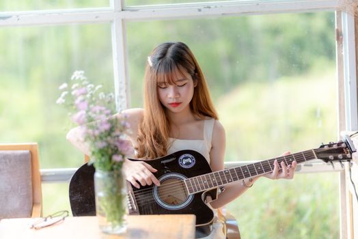 窗前，一位亚洲妇女弹着原声吉他，身旁放着鲜花，气氛明亮而宁静