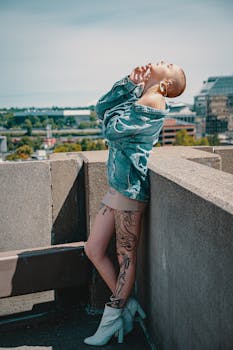 A fashion-forward woman with tattoos poses on a city rooftop in a denim jacket.