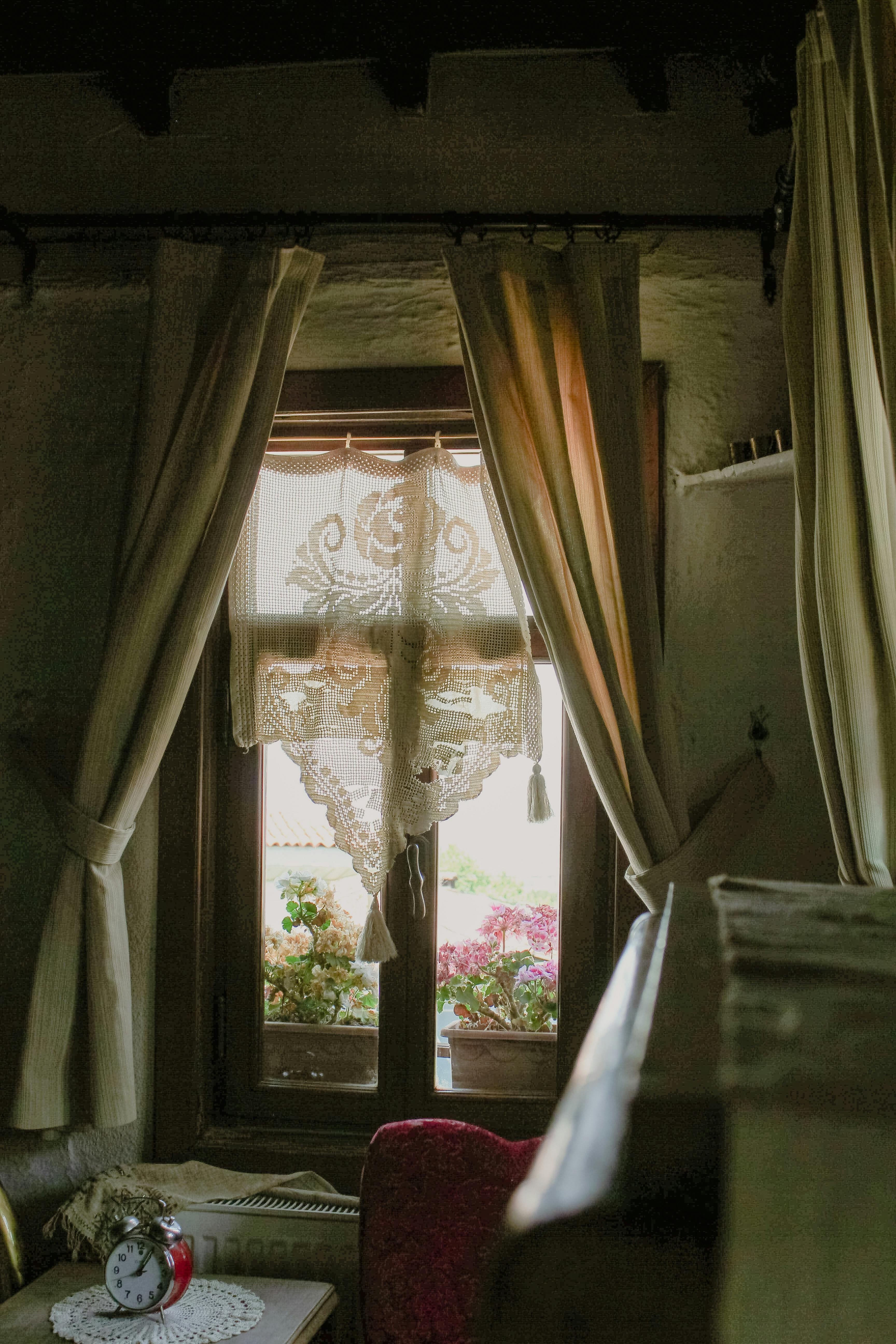 A cozy vintage interior with lace curtains framing a sunlit window and blooming flowers outside.