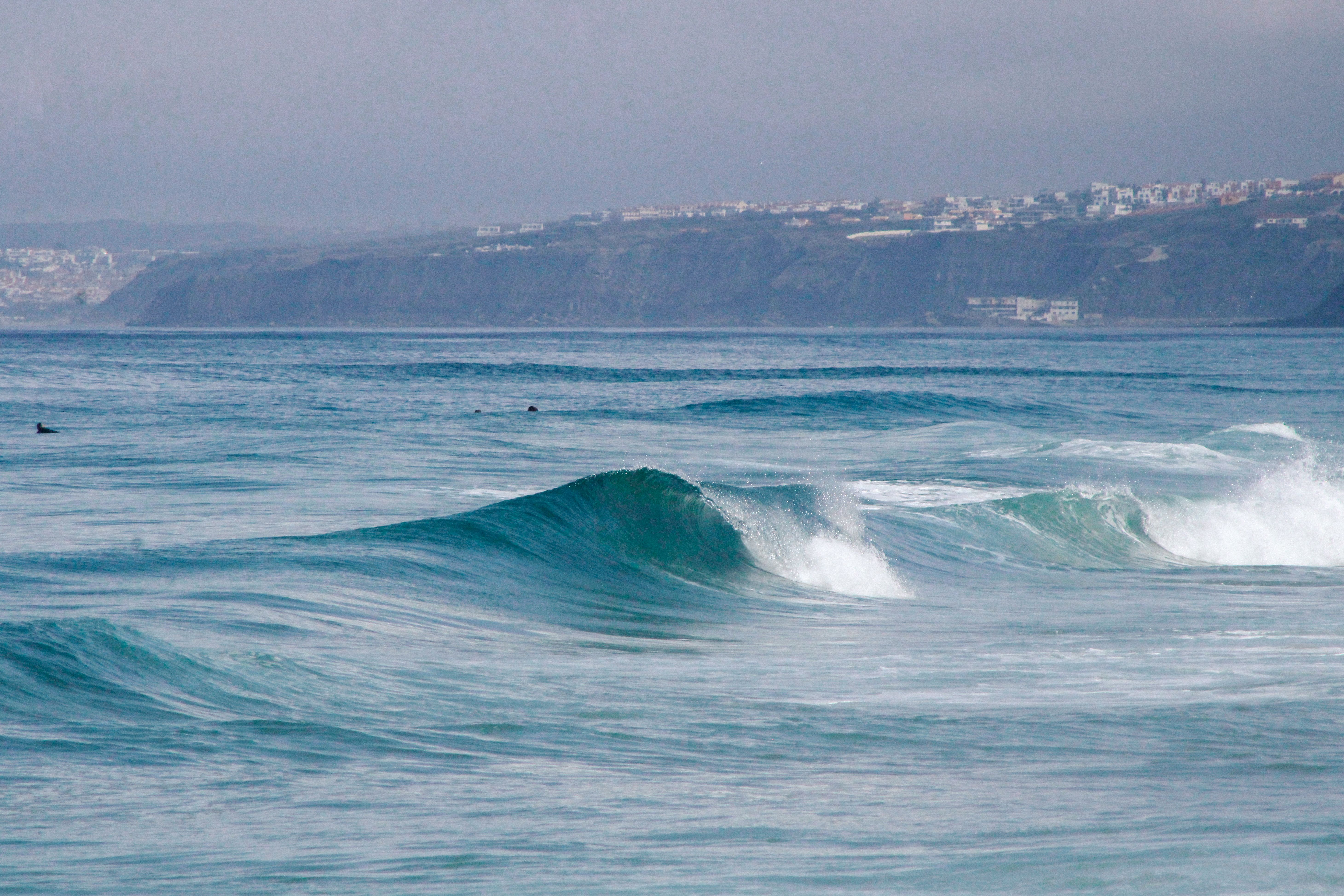 Vibrant ocean waves crashing against a rugged coastline with distant cliffs and town view.