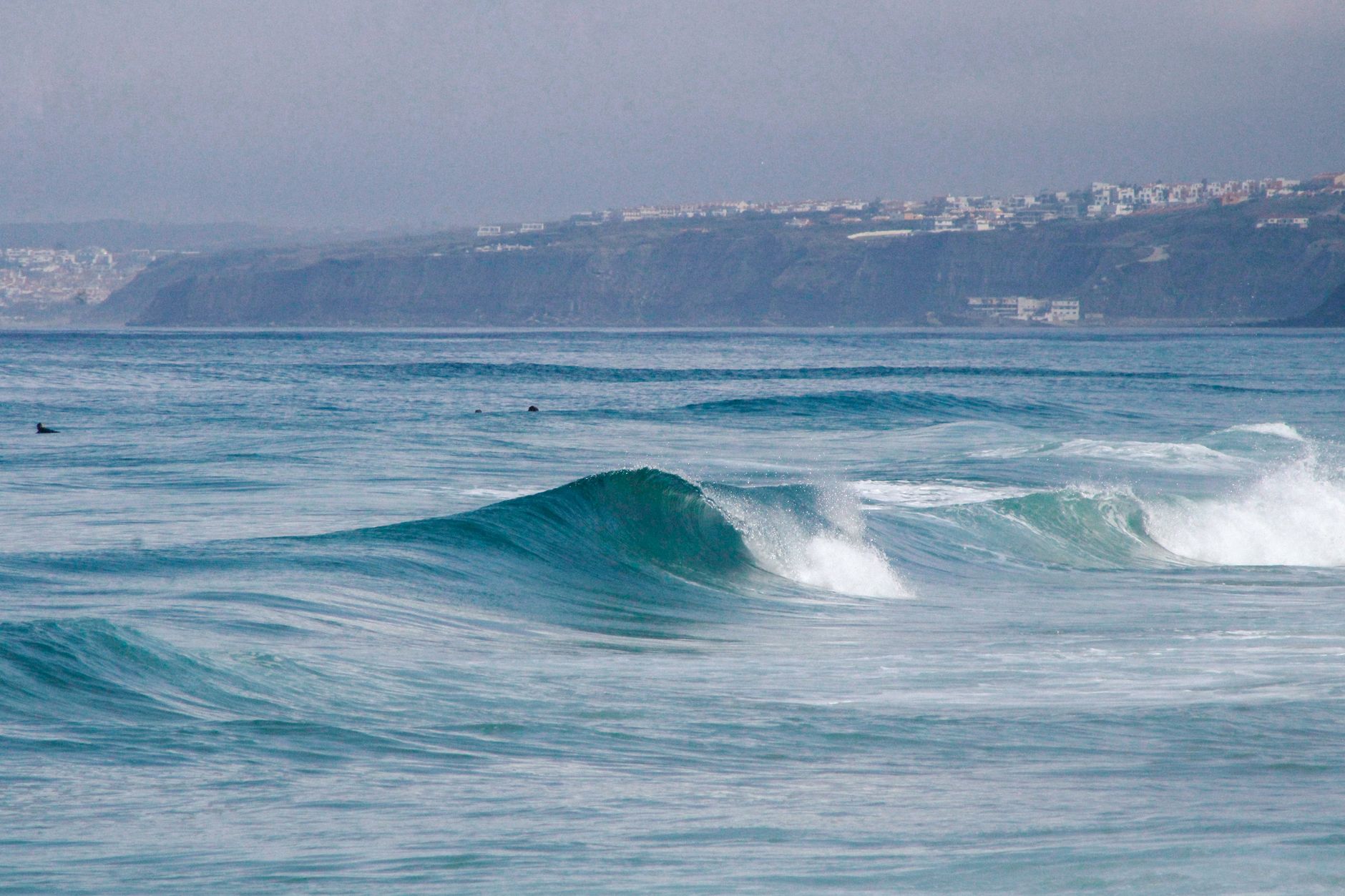 Vibrant ocean waves crashing against a rugged coastline with distant cliffs and town view.