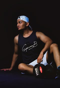Athletic man in white cap sitting indoors with a dumbbell, posing against a dark background.