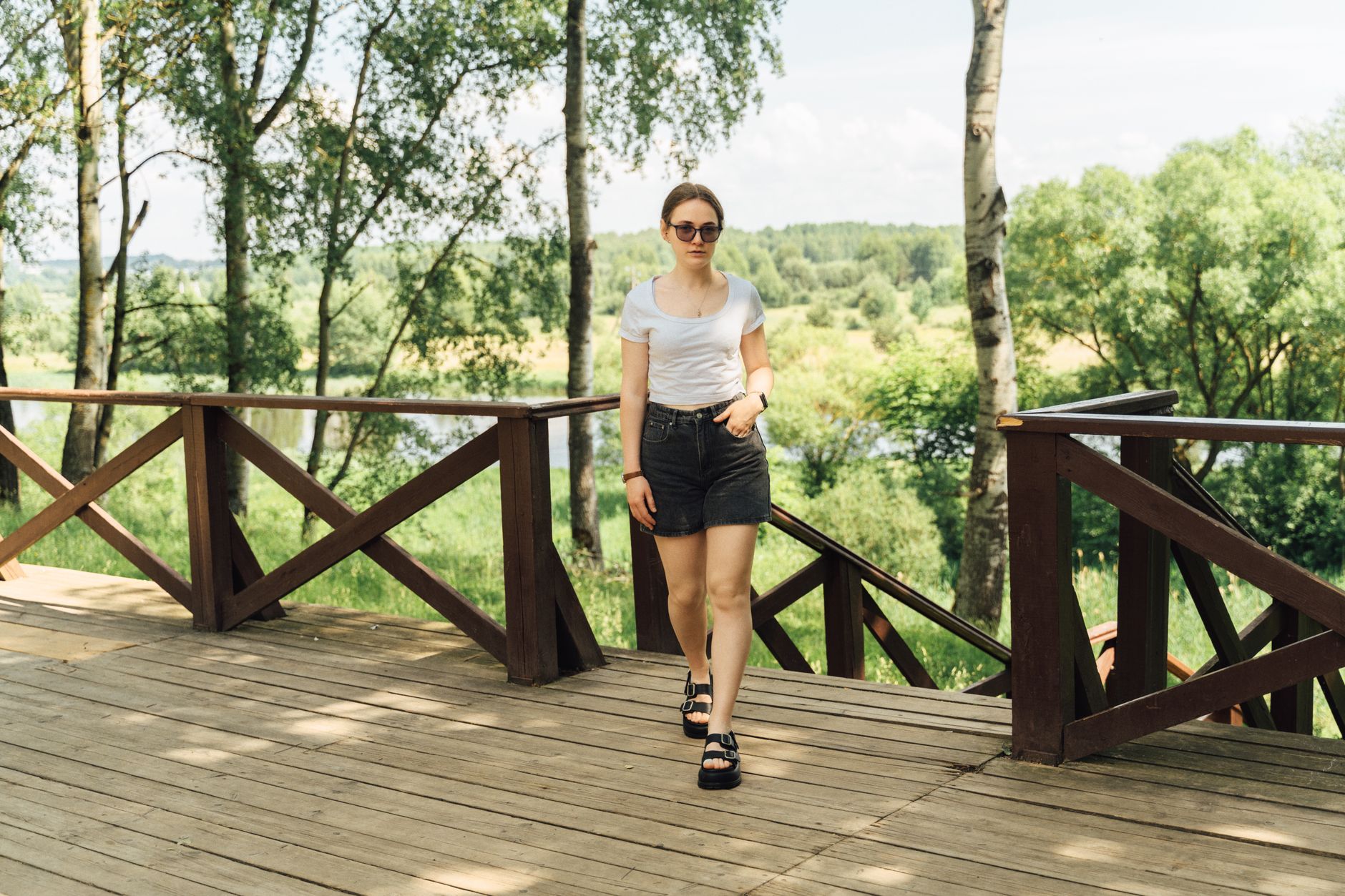 Casual summer fashion portrait of a woman walking on a wooden deck surrounded by lush greenery.