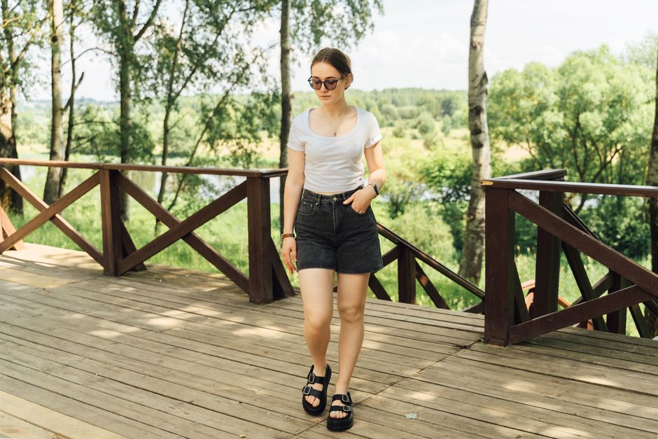 Woman in casual summer attire walks on a wooden deck amid lush green park.