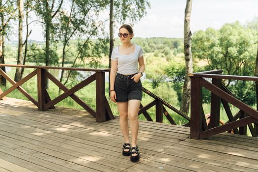 A woman in casual attire, walking on a wooden terrace during a sunny day in a rural setting.