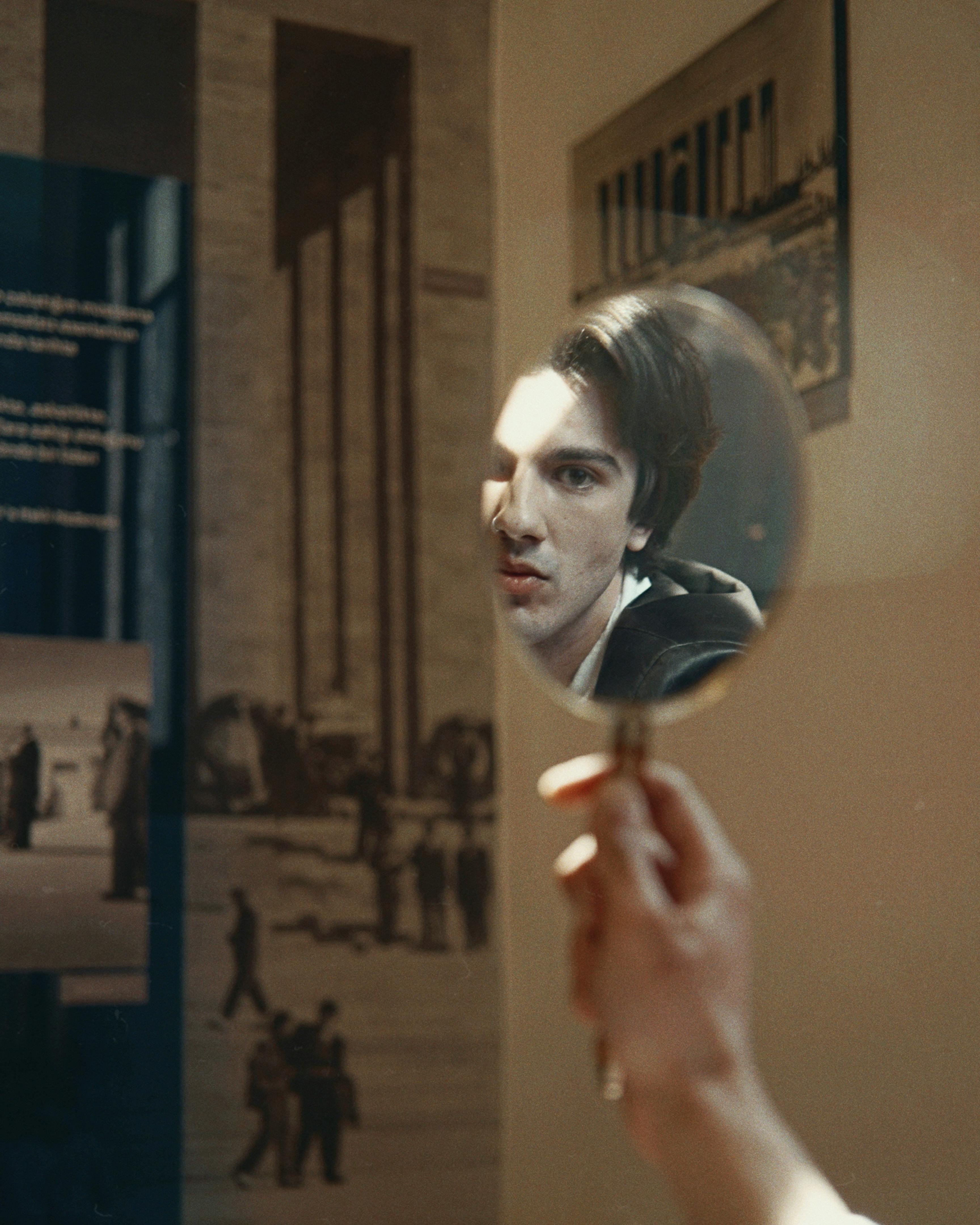 A man's face reflected in a handheld mirror inside with an artistic background.