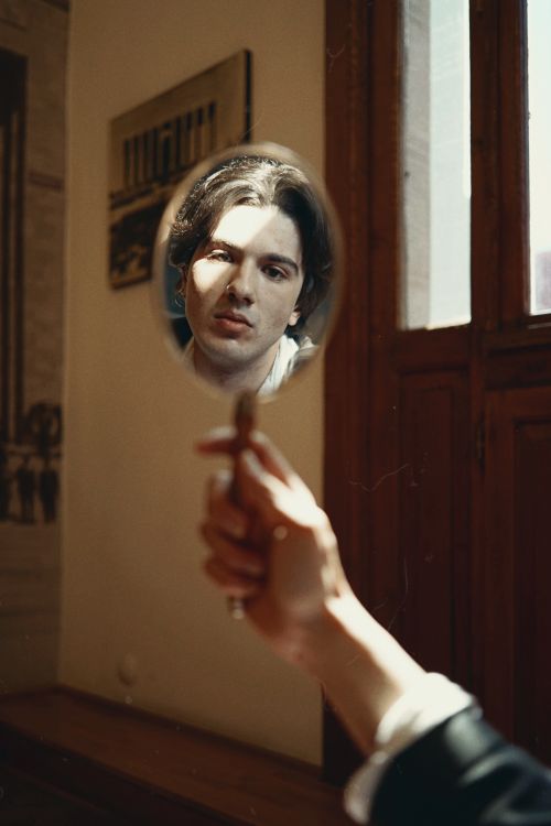 Free A young man gazes into a handheld mirror, creating a thoughtful reflection indoors. Stock Photo