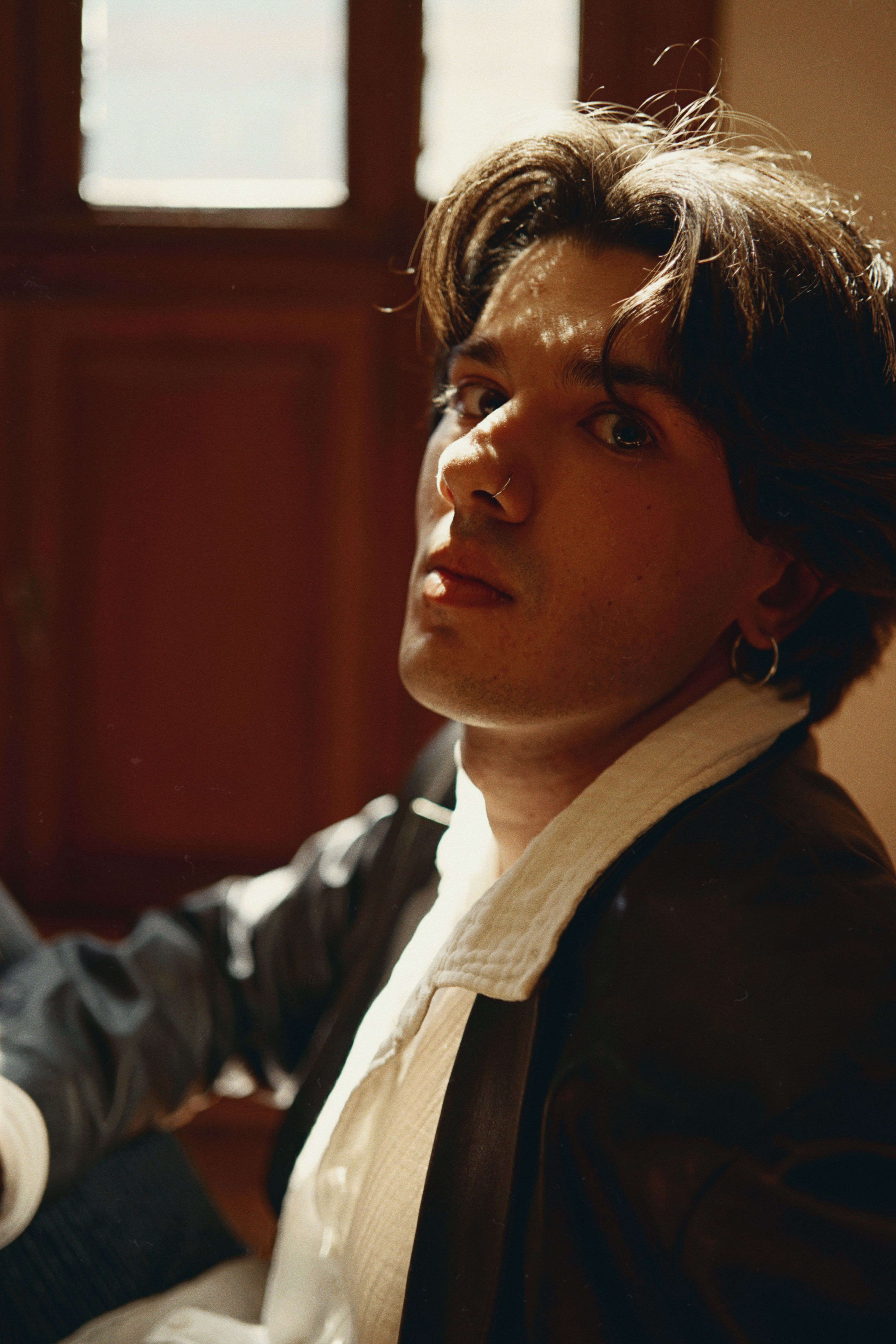 A young man with a white shirt and jacket poses indoors with sunlight creating a moody portrait.