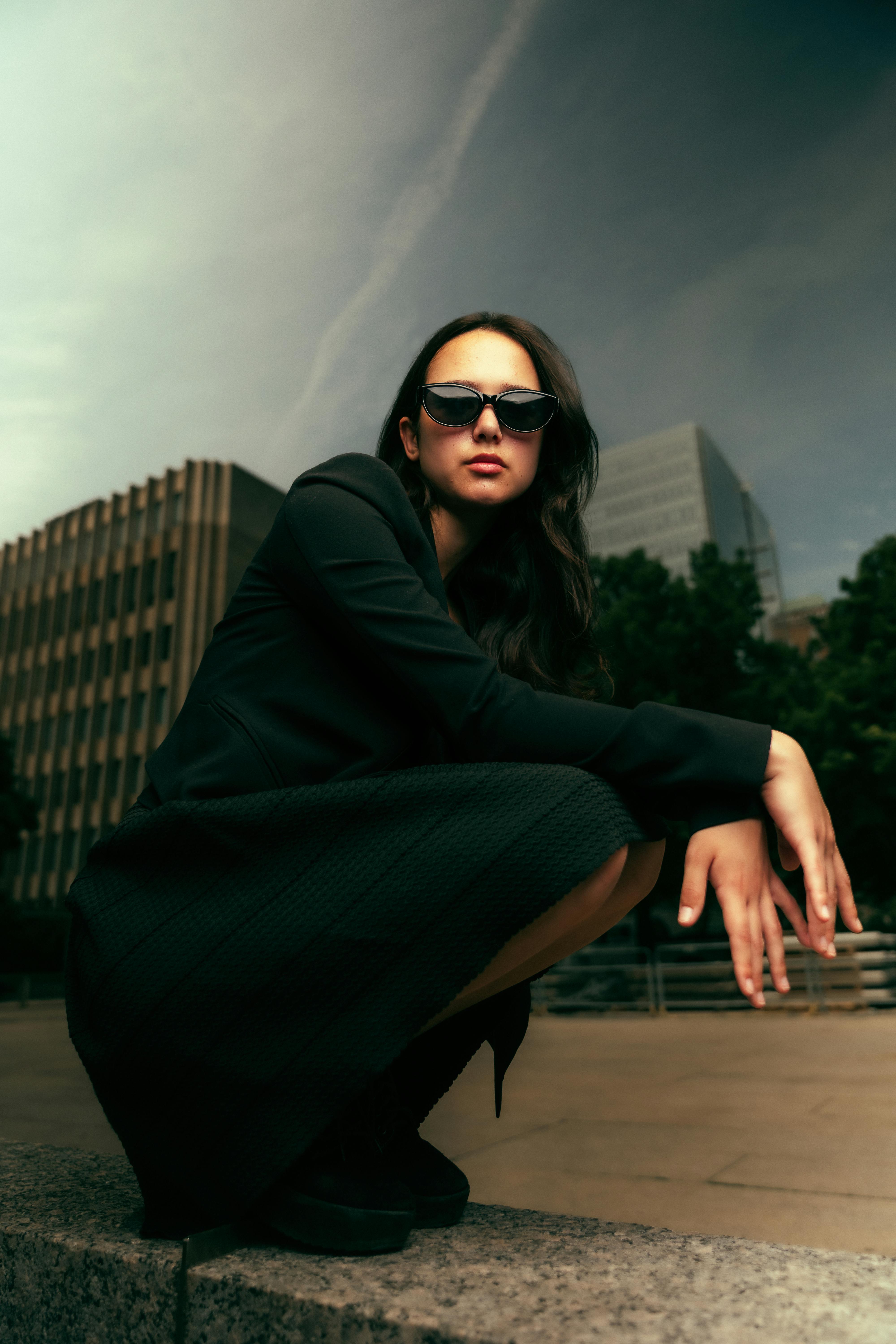 a woman in a black coat and sunglasses sitting on a ledge