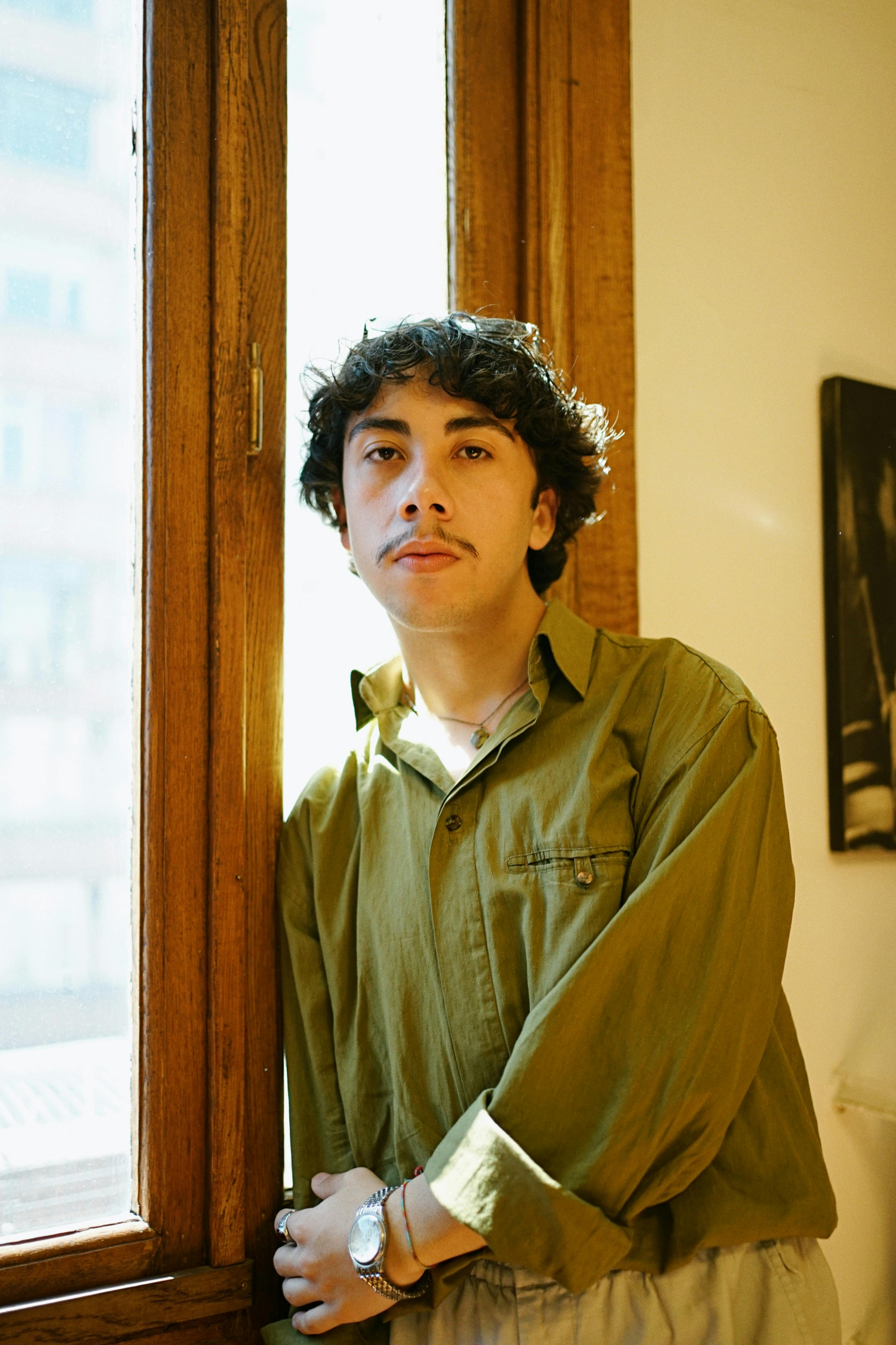 Portrait of a young man with curly hair standing indoors by a window in soft natural light.