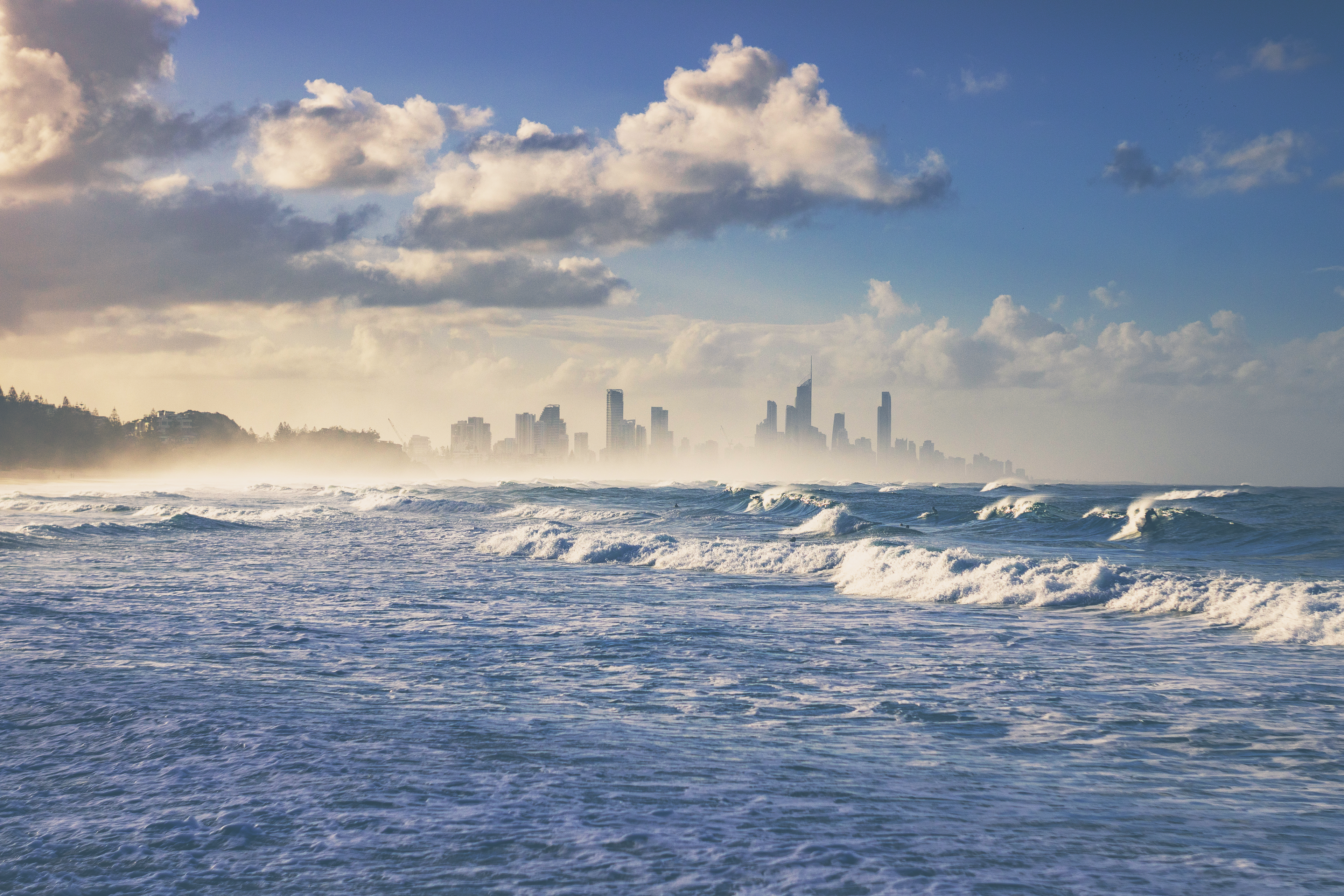 Beautiful ocean waves with the Gold Coast skyline in the background, under a partly cloudy sky.